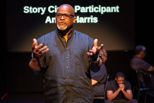 A man with glasses and a beard, wearing a dark shirt, speaking on a stage with a black background and text reading 'Story Circle Participant An...'
