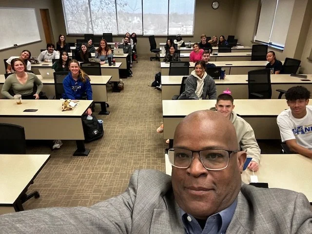 A classroom filled with students sitting at desks, with a teacher taking a selfie in the front. The classroom has large windows at the back, letting in natural light.