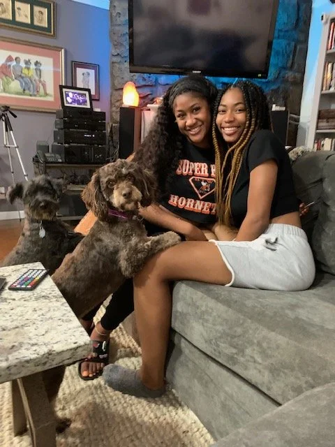 Two women sitting on a gray couch with two dogs, one of which is on the floor next to her. They are smiling and posing for the photo in a cozy living room. There is a TV on the wall behind them, and books and framed art on the walls.