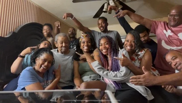 Group of smiling people celebrating indoors, some pointing and making gestures, with a drum in the background.