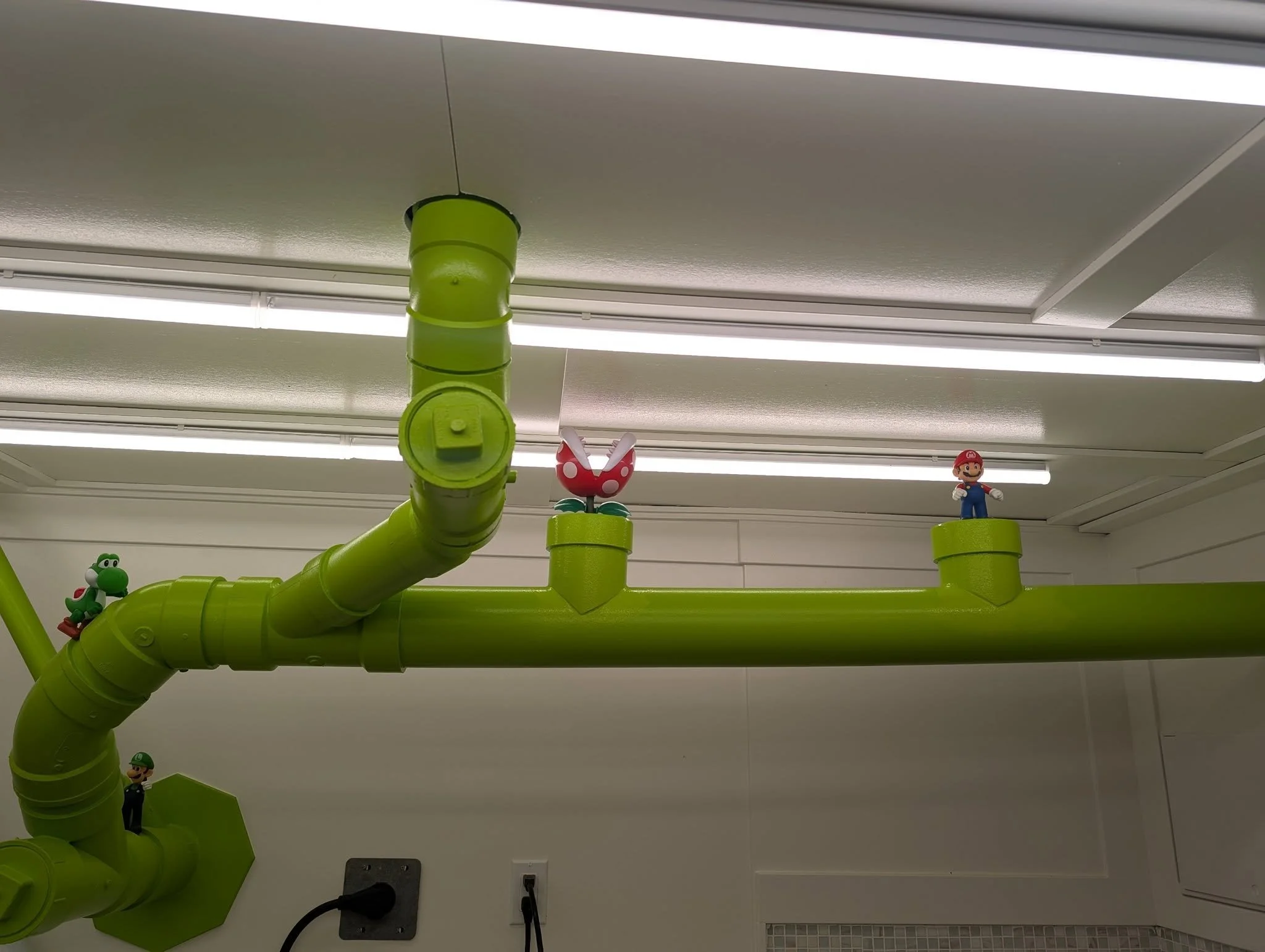 Green plastic pipes with figurines of Yoshi, Mario, and a Piranha Plant from Super Mario, set on the pipes in an indoor setting.