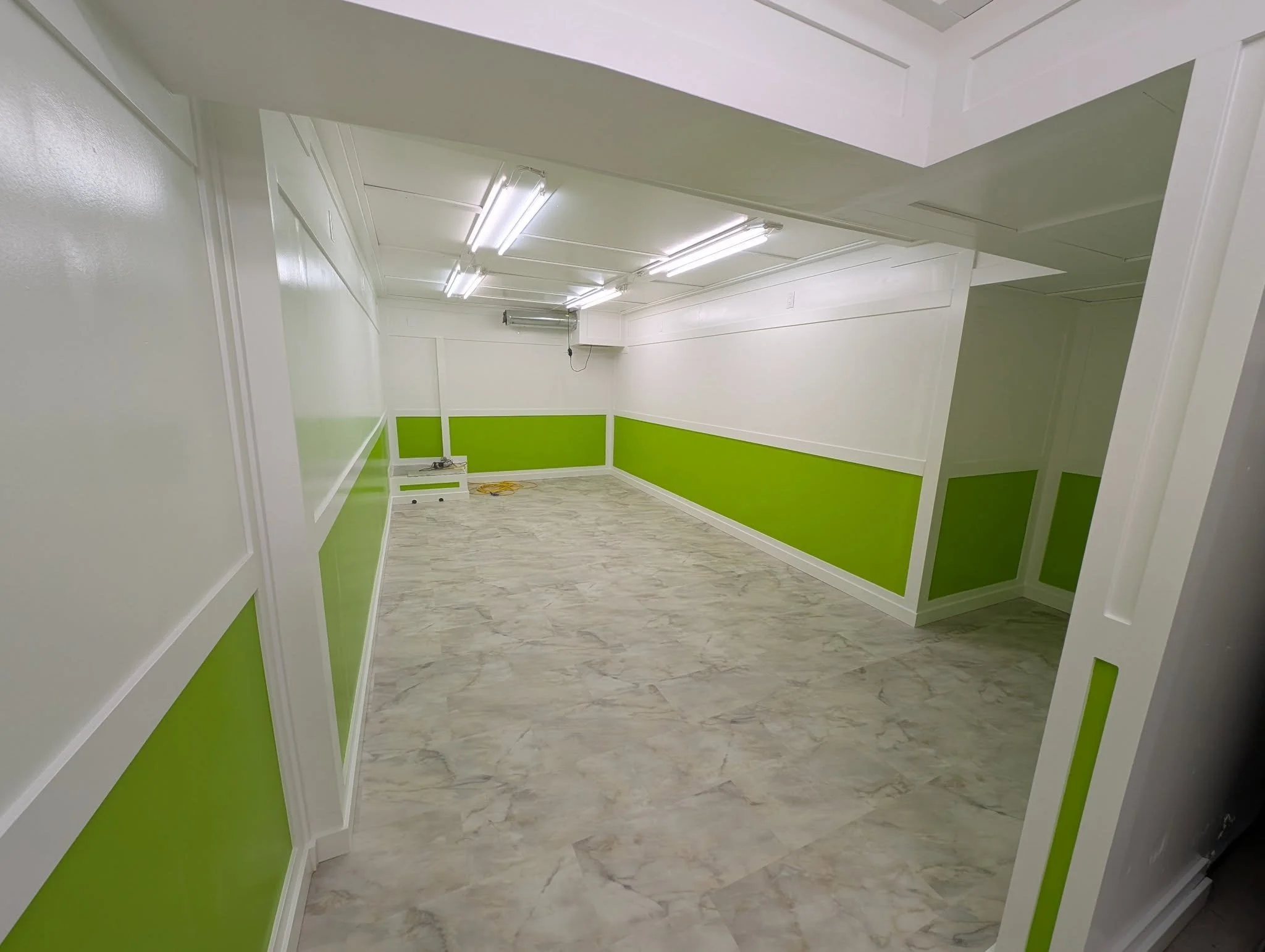 Empty room with white walls, green wainscoting, marble floor, fluorescent lighting, and a small window with an air conditioning unit.