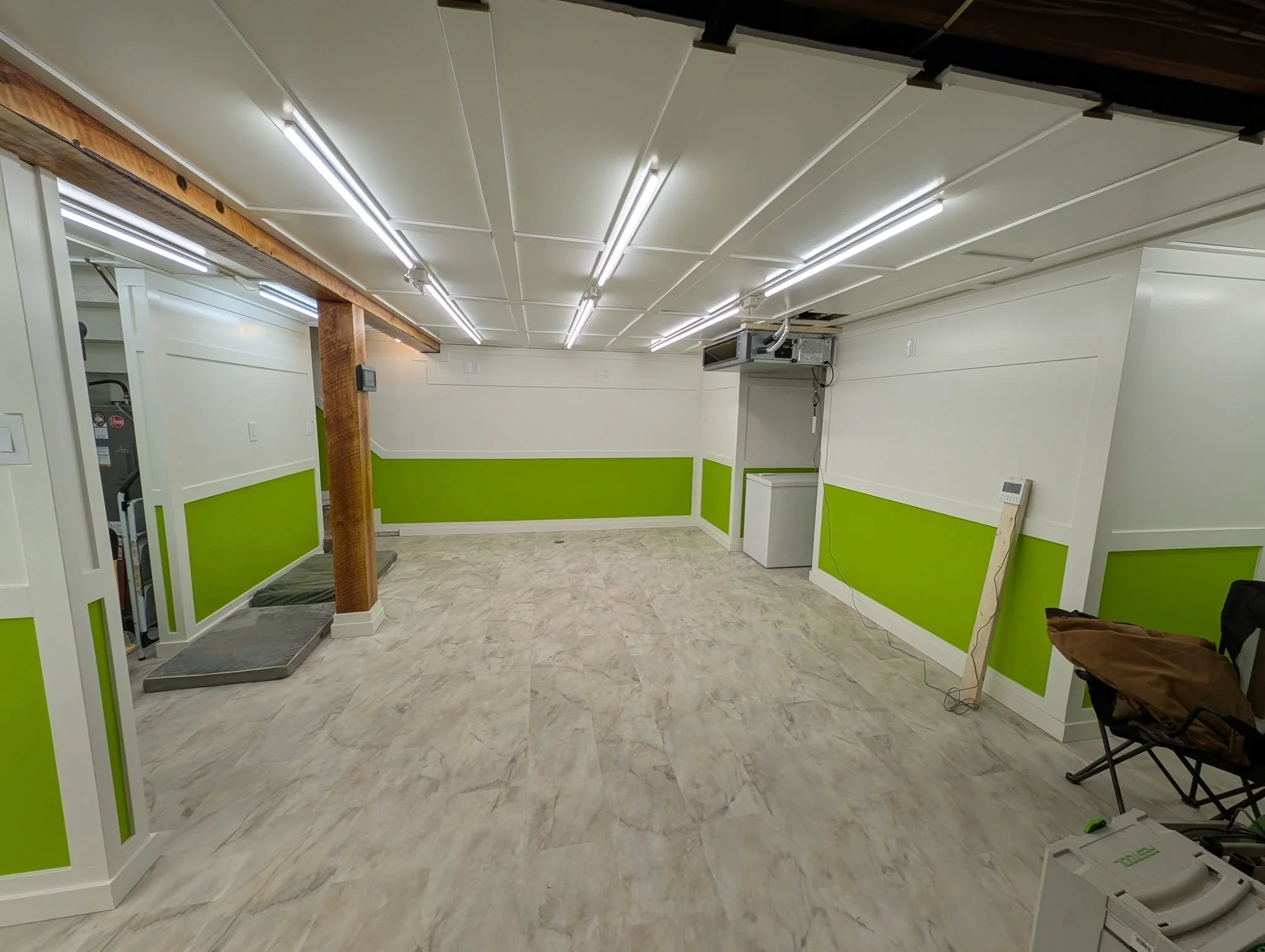 An empty room with white walls featuring a green wainscot, fluorescent lighting, and a wood support beam. There is some equipment and a chair on the right side, and a small white machine on the floor.