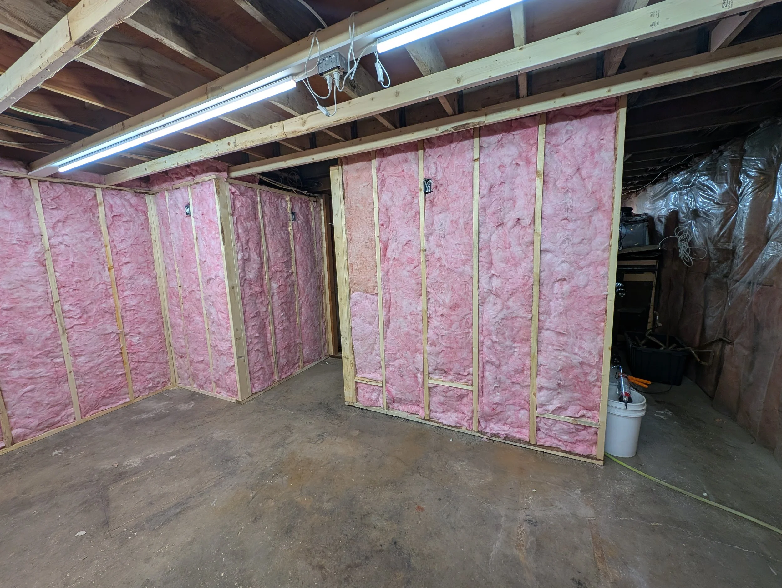 Construction of interior basement wall with pink insulation and wooden framing, exposed ceiling with fluorescent lighting, and construction tools and supplies in the corner.