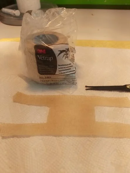 A roll of 3M Veltrim tape in clear plastic packaging, a pair of scissors, and two brown tape strips on a surface.
