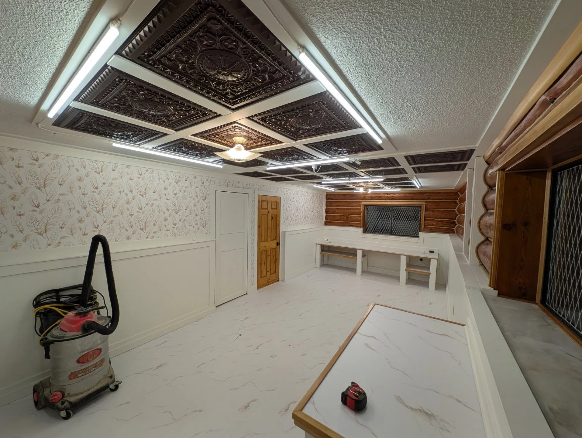 Room with decorative ceiling panels, wallpaper with leaf pattern, wooden doors, and workbench with tools, possibly under renovation.