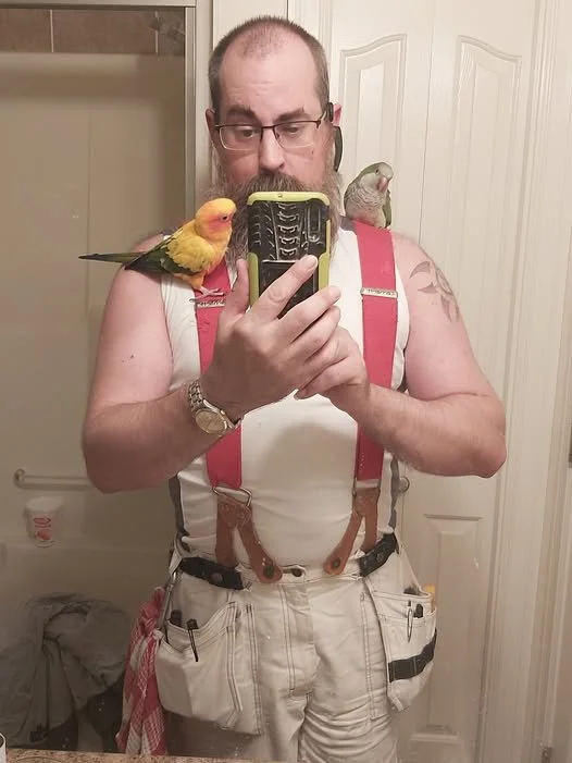 Mirror selfie of a man with a beanie, glasses, and suspenders, taking a photo with two parrots on his shoulders in a bathroom.