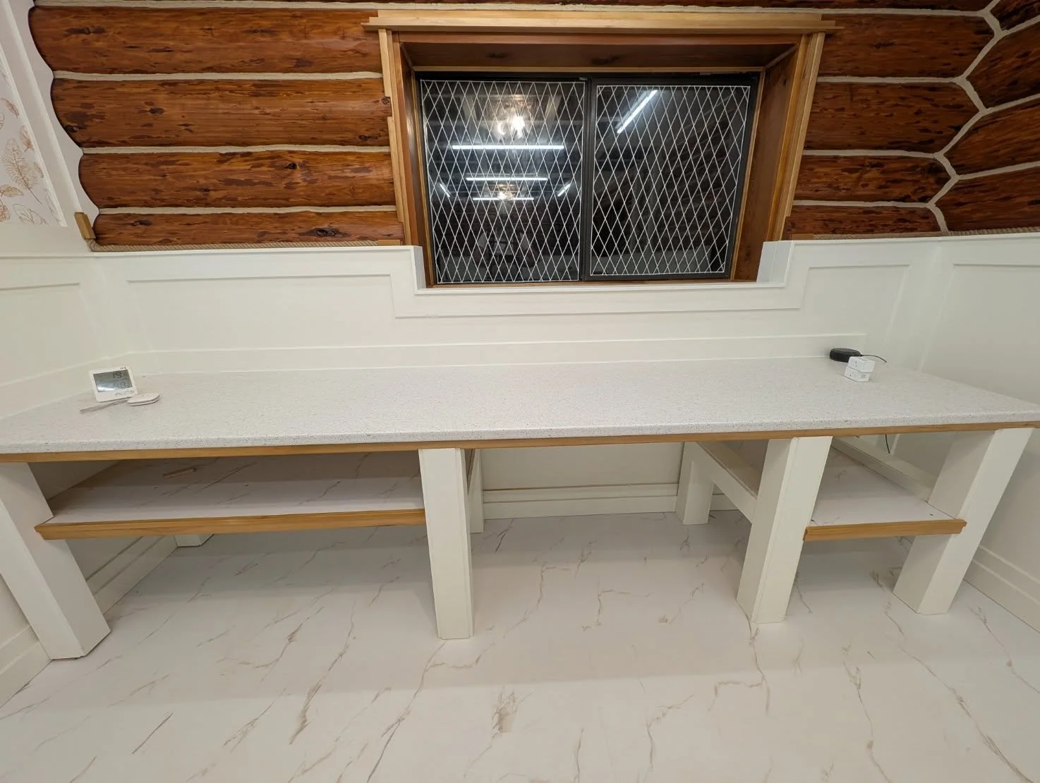 White countertop with shelving beneath, a window above, and a wooden wall behind. The countertop has a thermostat on the left, an electronic device on the right, and a power adapter. The floor is marble or marble-like tile.