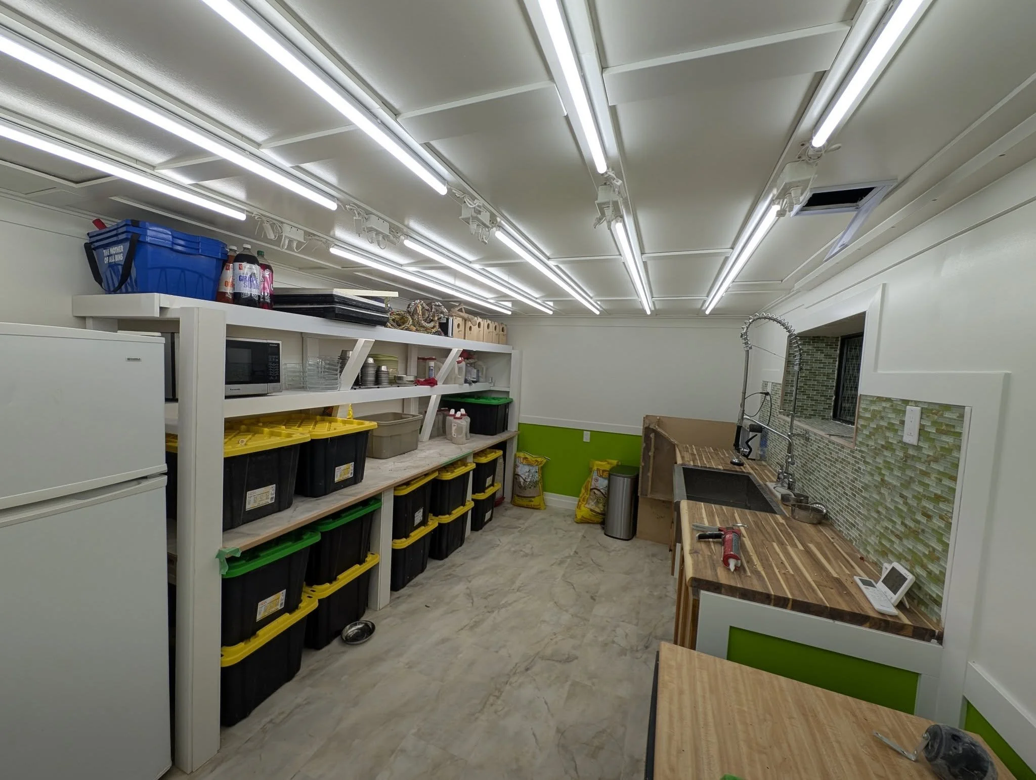 A well-lit kitchen or utility room with white walls and ceiling, wood countertops, a sink with a tall, flexible faucet, and a mosaic tile backsplash. There are shelving units with storage bins, a microwave, a refrigerator, and various cleaning suppli