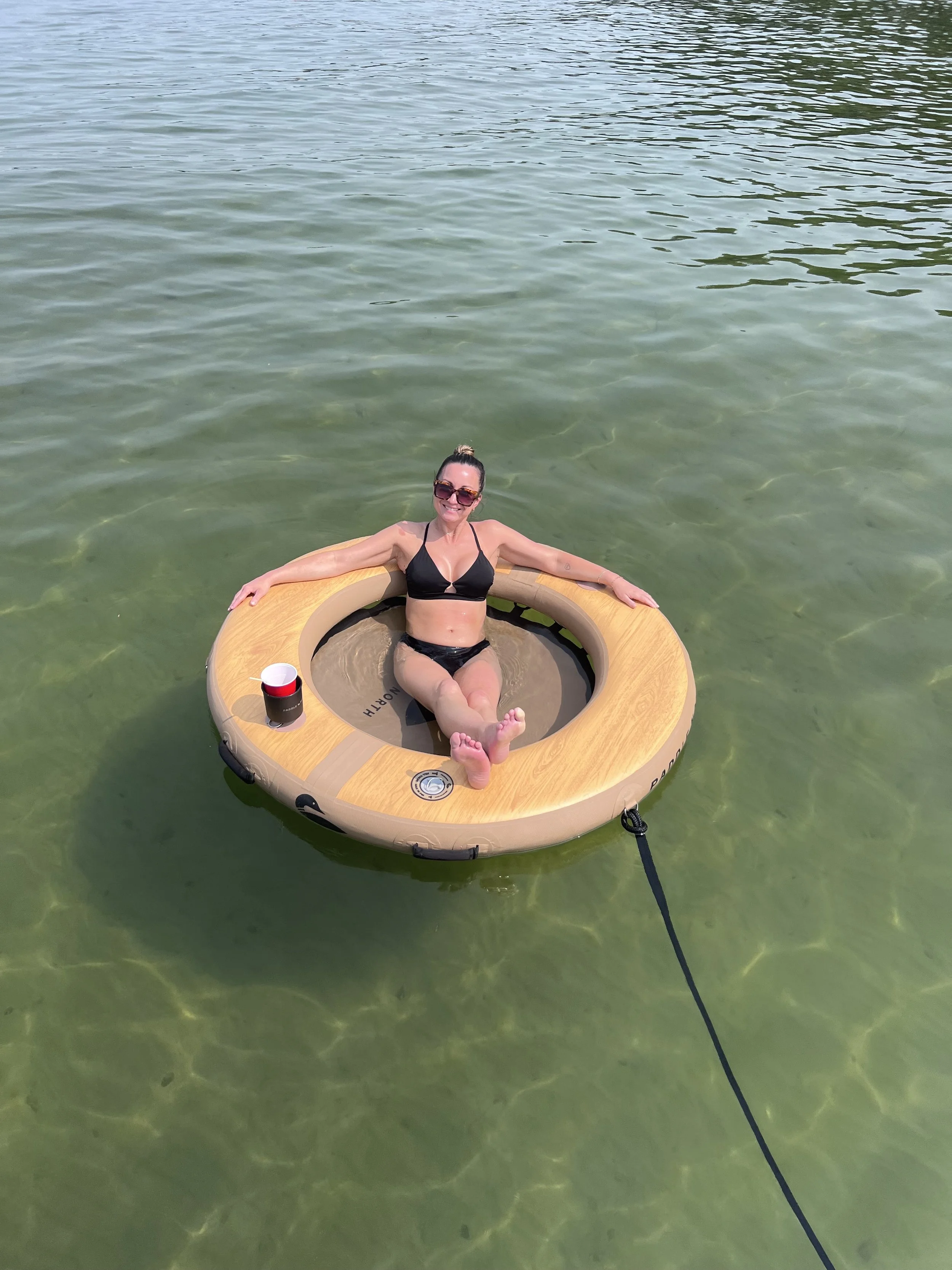 A woman relaxing on a round float in the water, wearing a black bikini, sunglasses, and smiling. There is a red cup on the float.