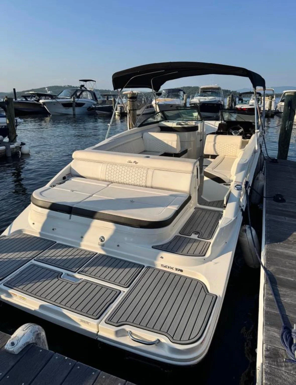 A white motorboat docked at a marina, with cushioned seating area at the front, a covered steering area, and other boats visible in the background on the water.