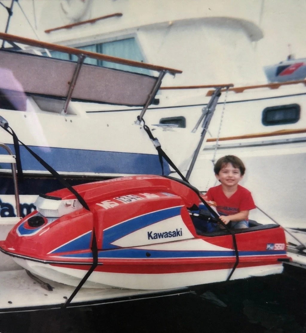 A young boy in a red shirt sitting inside a red, white, and blue Kawasaki jet ski docked at a marina, with a large white boat in the background.