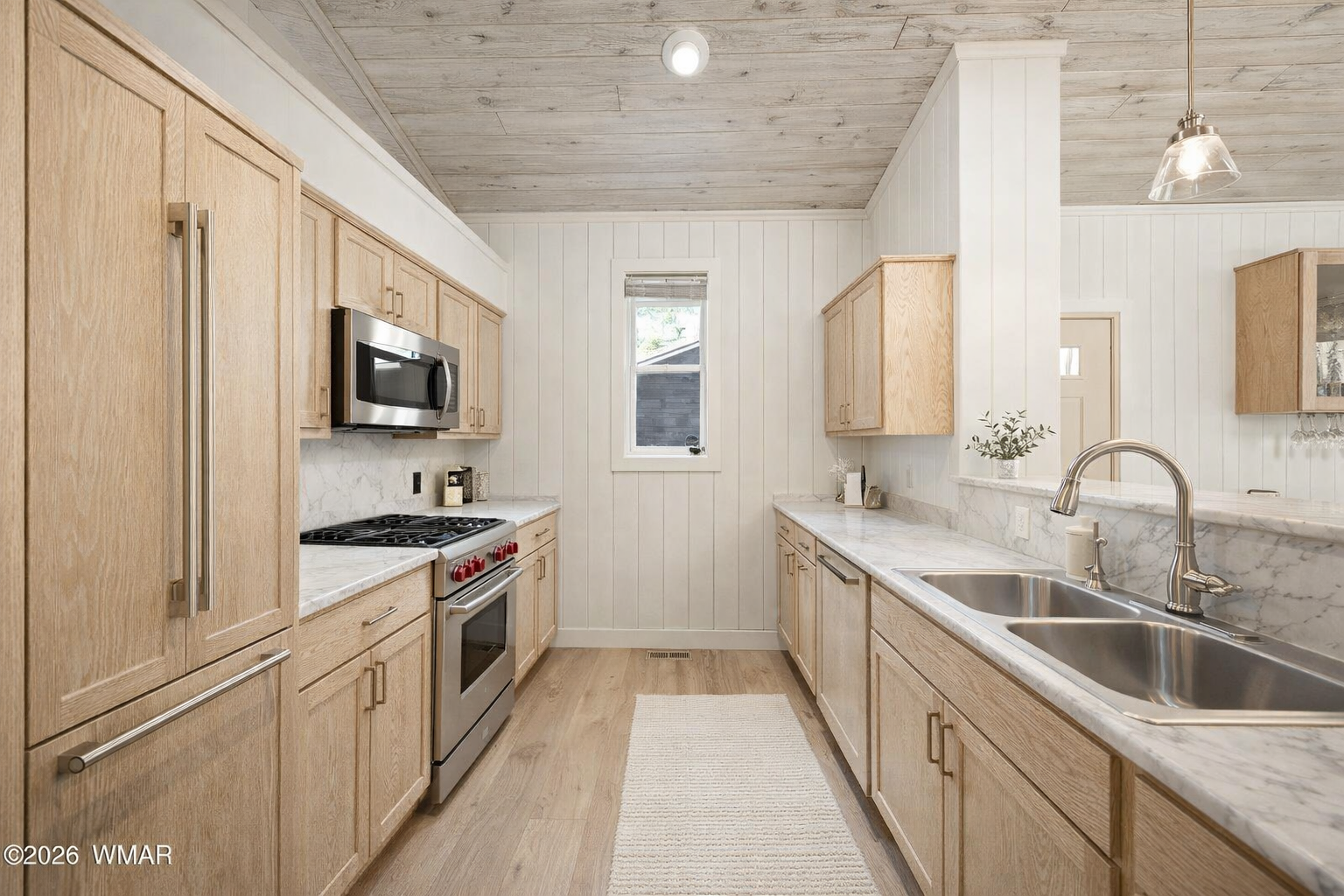 Kitchen with light wood cabinets, marble countertops, stainless steel sink, stove, microwave, and small window, with a pendant light and white wall paneling.