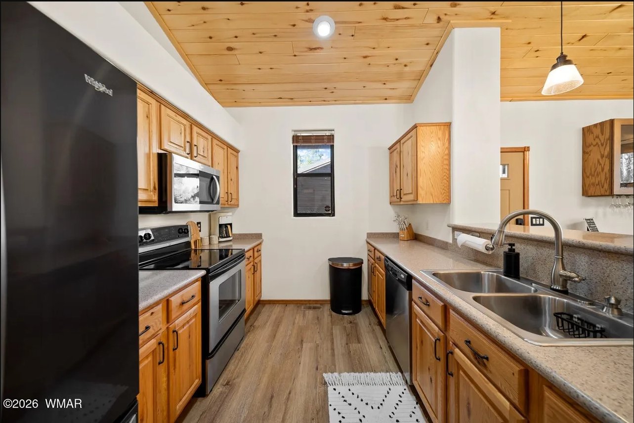 A kitchen with wooden cabinets, stainless steel appliances, a double sink, and a granite countertop. There is a small window, a trash can, and hanging light fixtures.