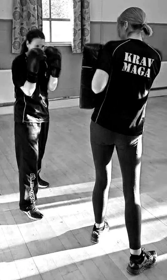 Two women in sports attire practicing boxing in a gym. One woman is wearing gloves and a black shirt, while the other, standing with her back to the camera, has