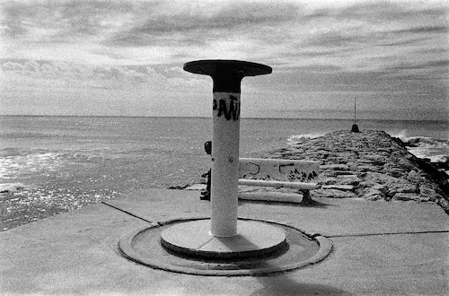 A black-and-white photo of a harbor with a concrete pier extending into the water, featuring a round pedestal with a thick top at the end.