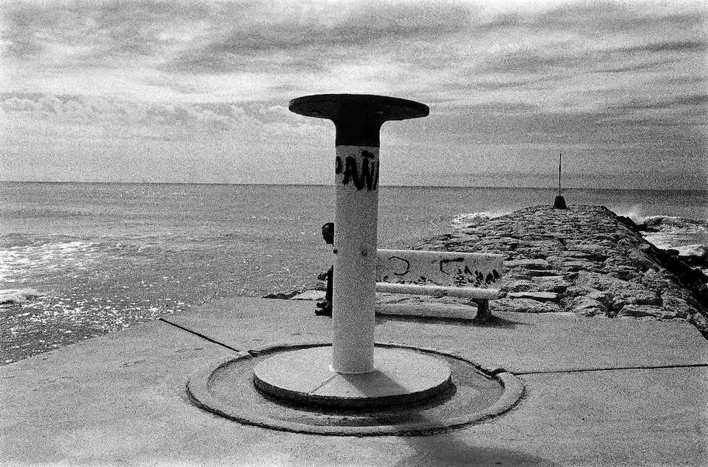 Melted Horizon - Black and white minimalist photograph of a white column overlapping a stone bench by the sea, with a blurred figure subtly merging into the structure. From the series One.