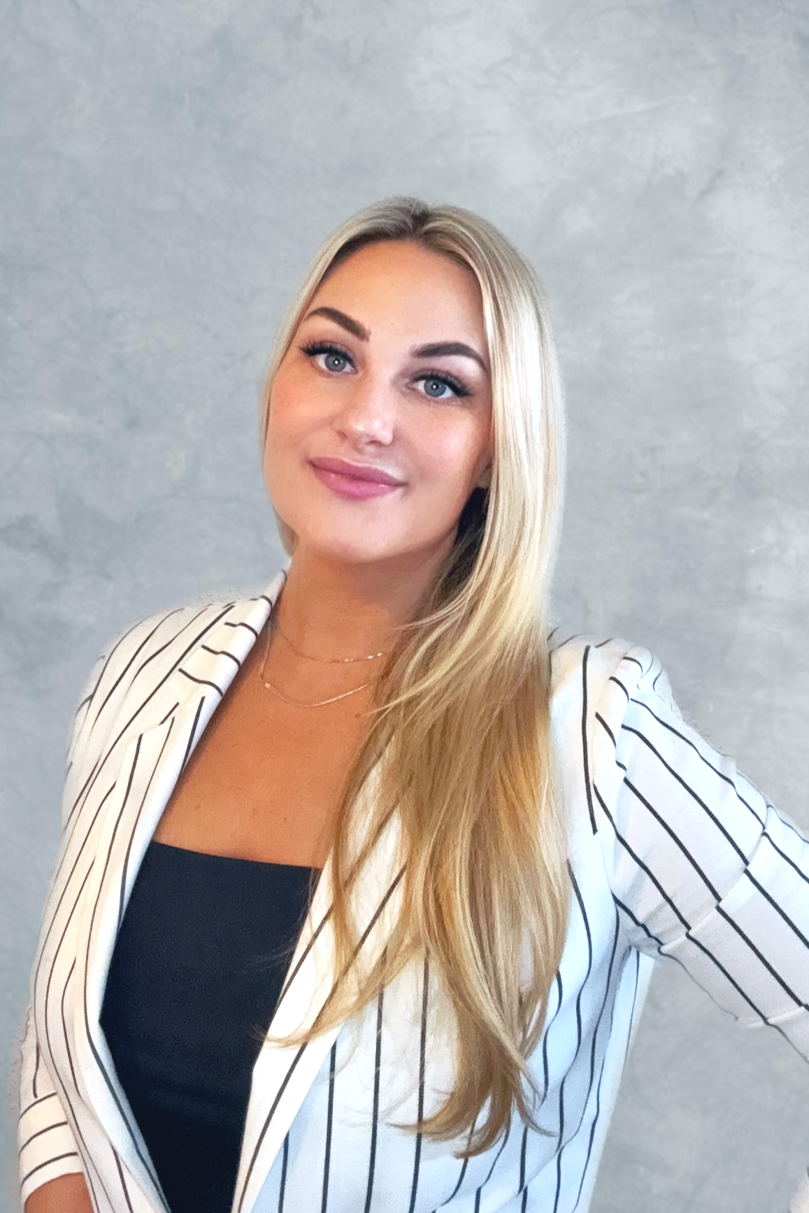 Portrait of a young woman with long blonde hair, blue eyes, arched eyebrows, wearing a striped blazer over a black top, smiling, against a neutral gray background.
