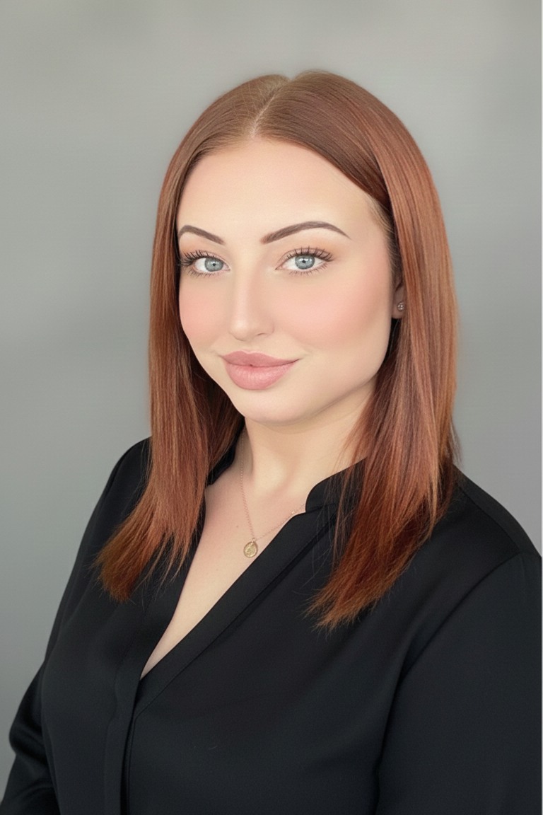 A young woman with straight red hair, light blue eyes, and light skin, wearing a black blouse and a gold necklace, looking at the camera with a slight smile, against a plain light gray background.