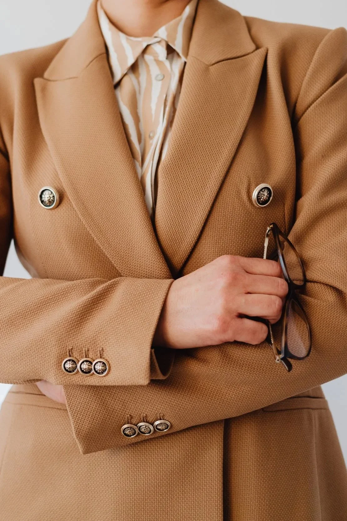 Person wearing a tan blazer with patterned buttons, holding glasses in hand and wearing a striped shirt underneath.