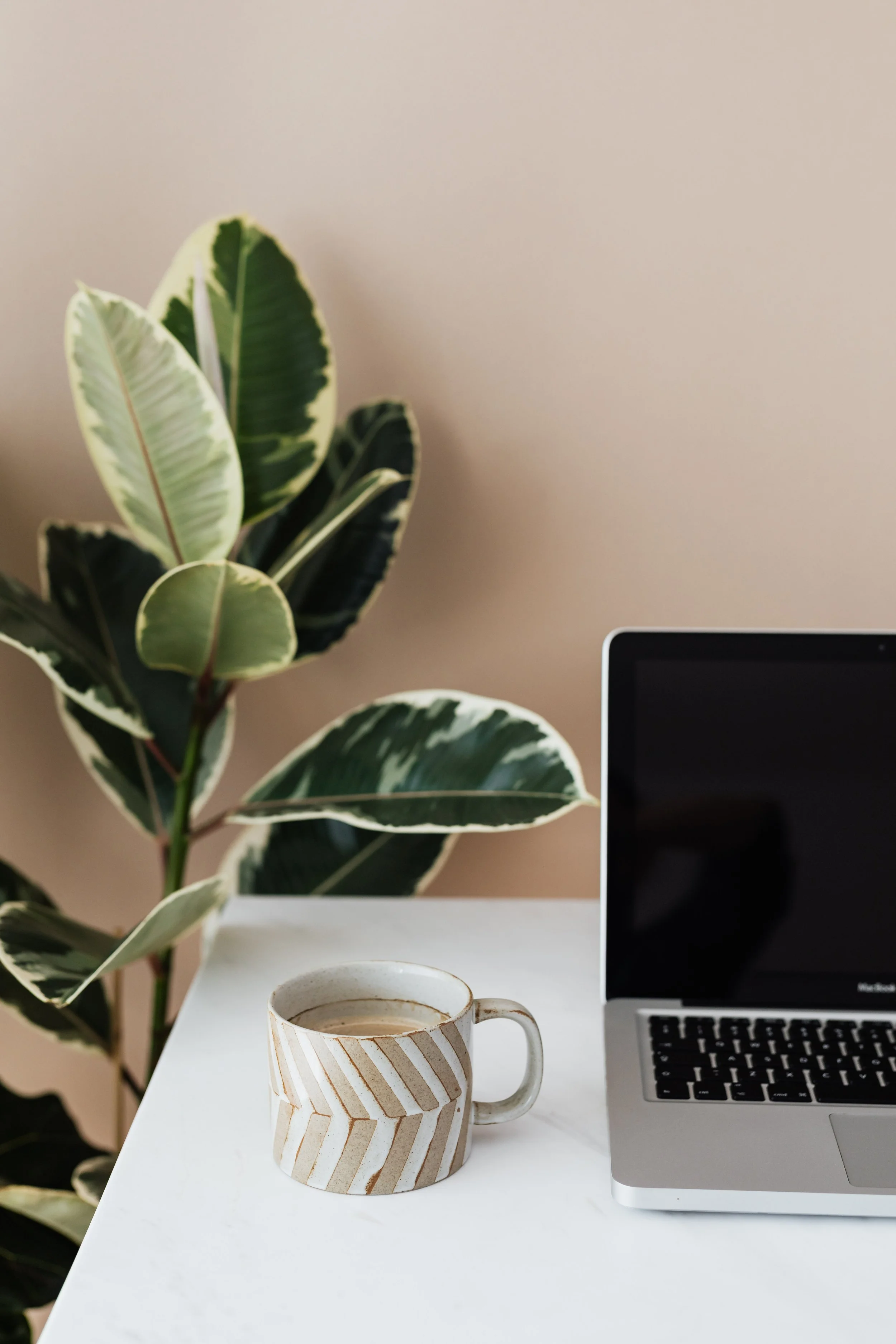 A white table with a patterned mug filled with coffee, an open laptop, a potted plant with variegated leaves, and a ceramic cup, against a beige wall.