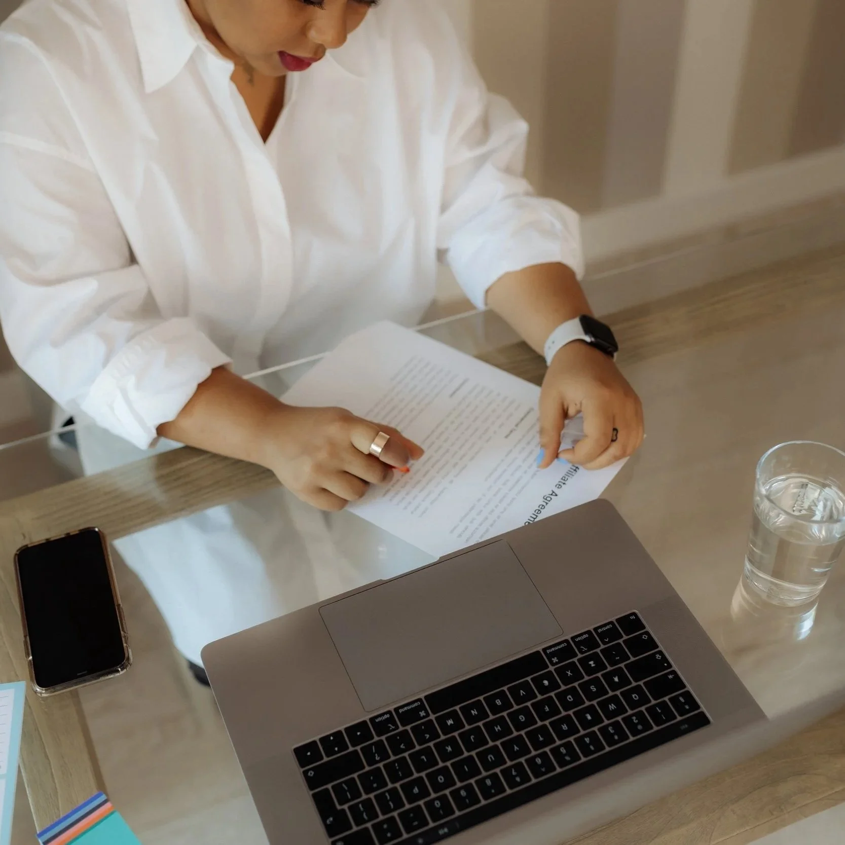 A woman in a white shirt sitting at a desk with a silver laptop, a smartphone, a glass of water, and colorful sticky notes.