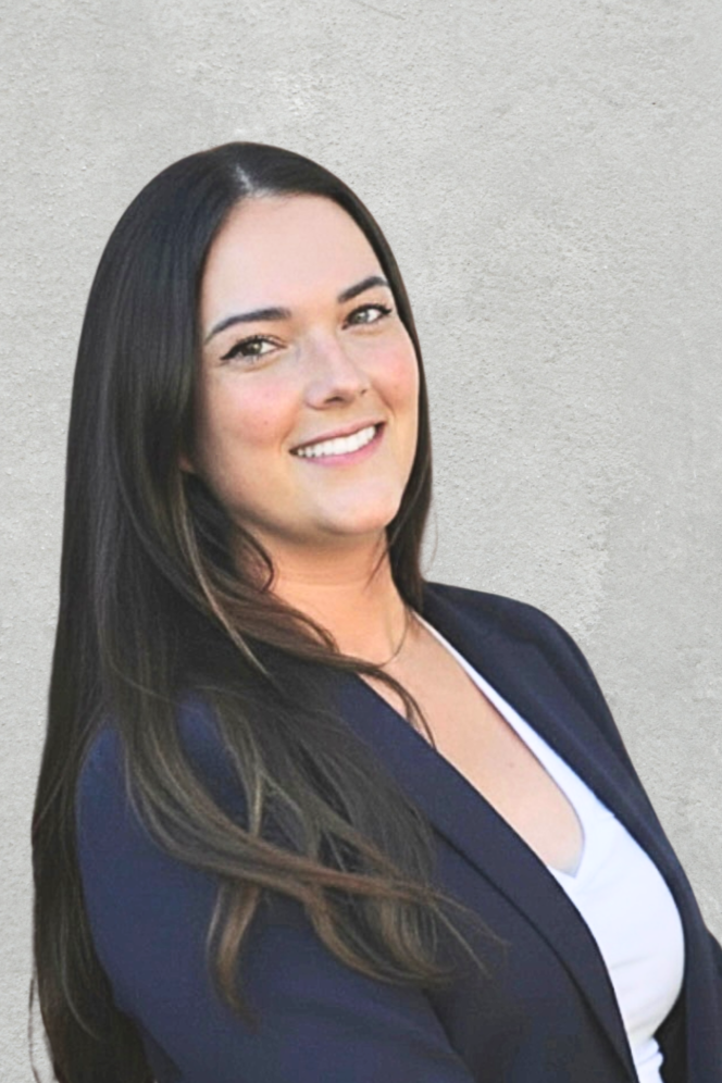 A woman with long dark hair smiling, wearing a navy blazer over a white top, standing against a light gray wall.