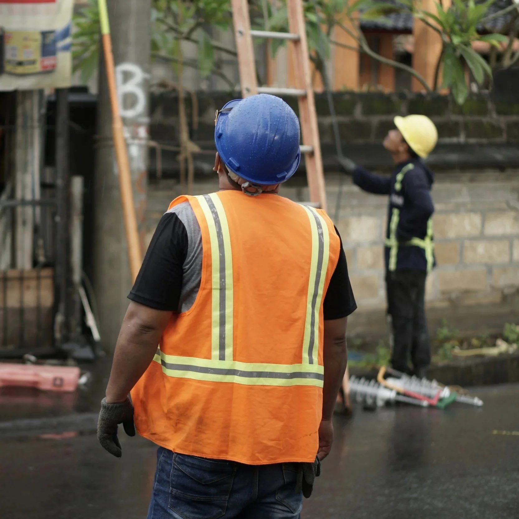 Construction worker wearing an orange safety vest and a blue helmet, standing on a construction site, looking towards another worker wearing a yellow helmet and black uniform, with construction materials and tools in the background.