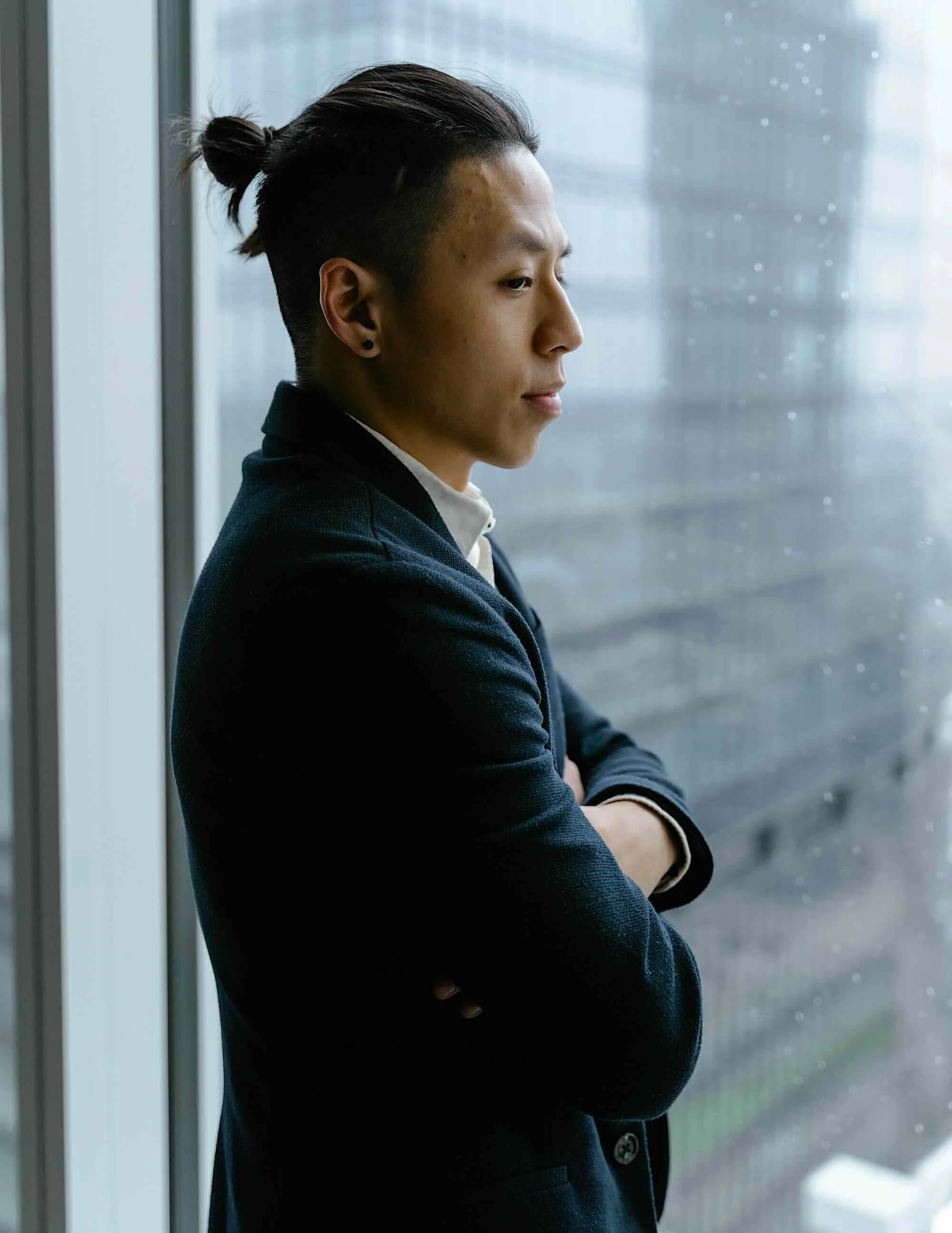 A young man with short hair tied back, wearing a dark blazer and white shirt, standing with arms crossed and looking out a rain-streaked window.