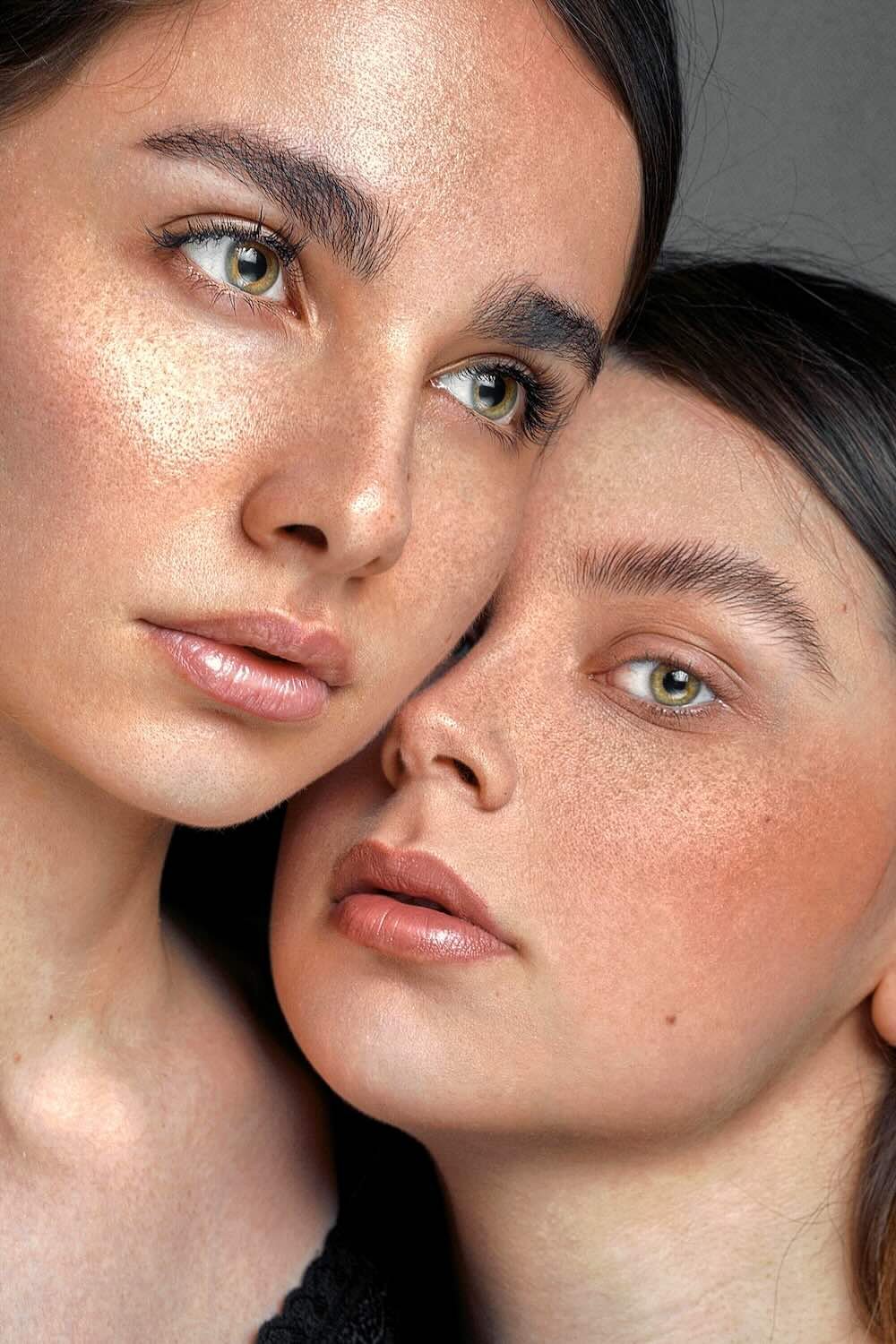 Close-up of two women with natural makeup and visible freckles, touching faces and looking into the camera.