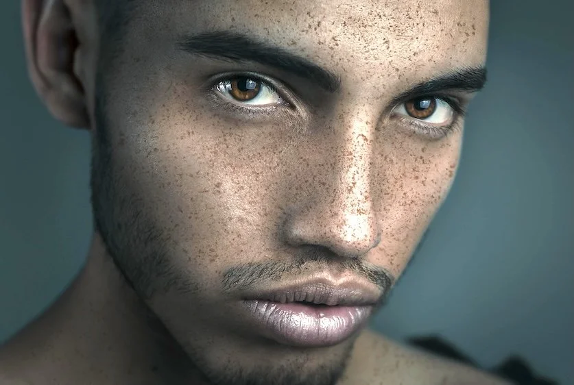 Close-up portrait of a young man with brown eyes, freckles, and short facial hair, looking intensely at the camera.