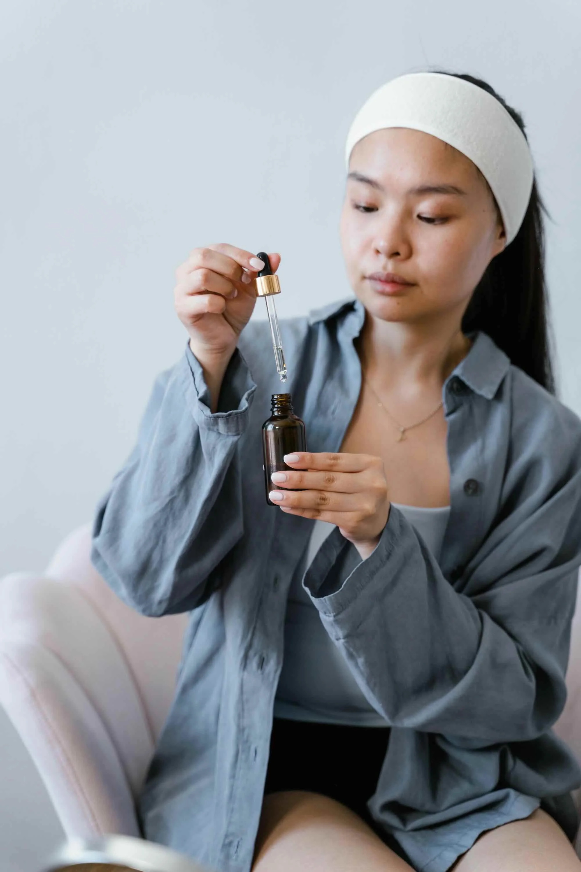 A woman wearing a white headband and gray shirt is using a dropper to transfer something from a dark glass bottle.