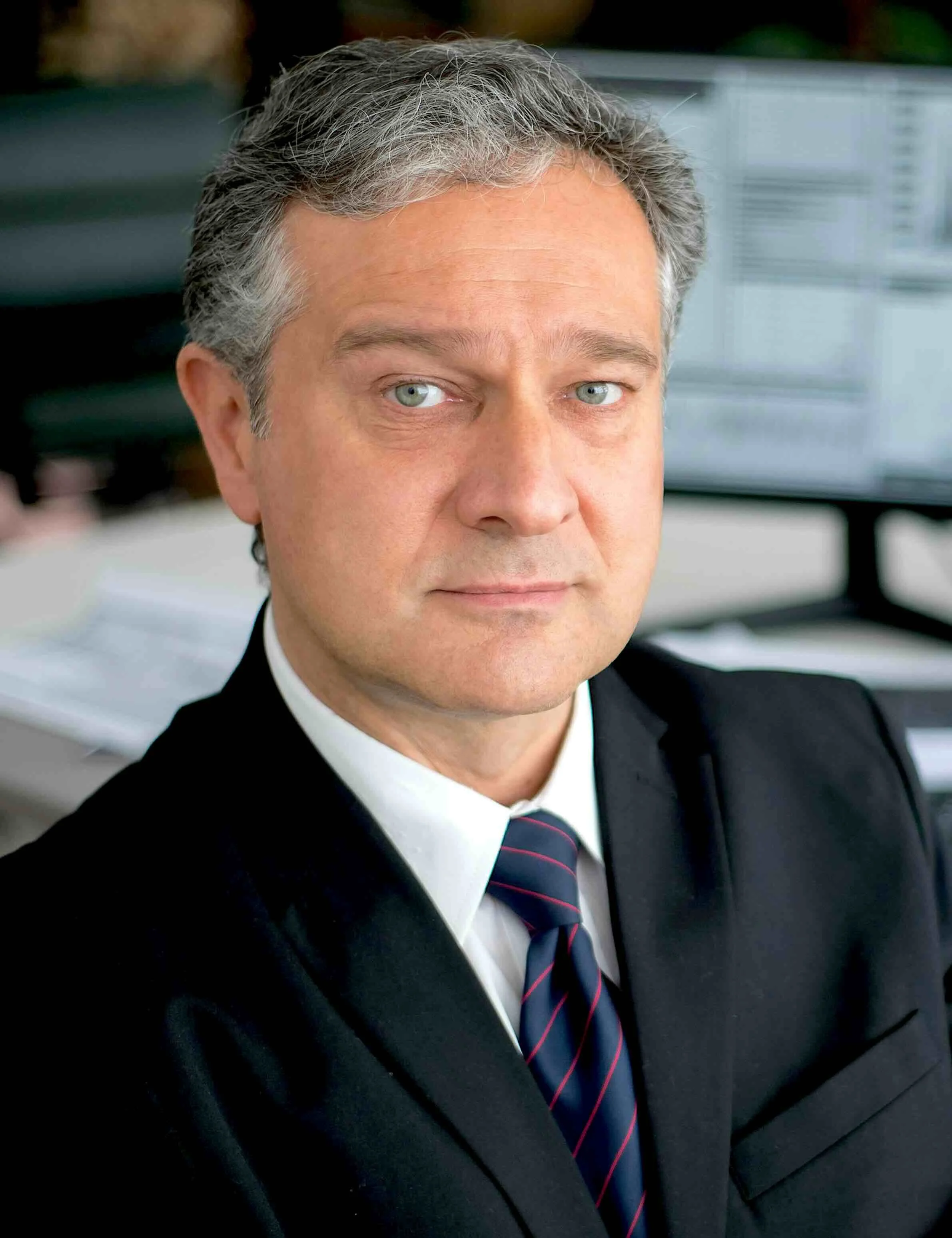 A middle-aged man with gray hair and blue eyes wearing a black suit, white shirt, and striped tie sitting in an office environment.