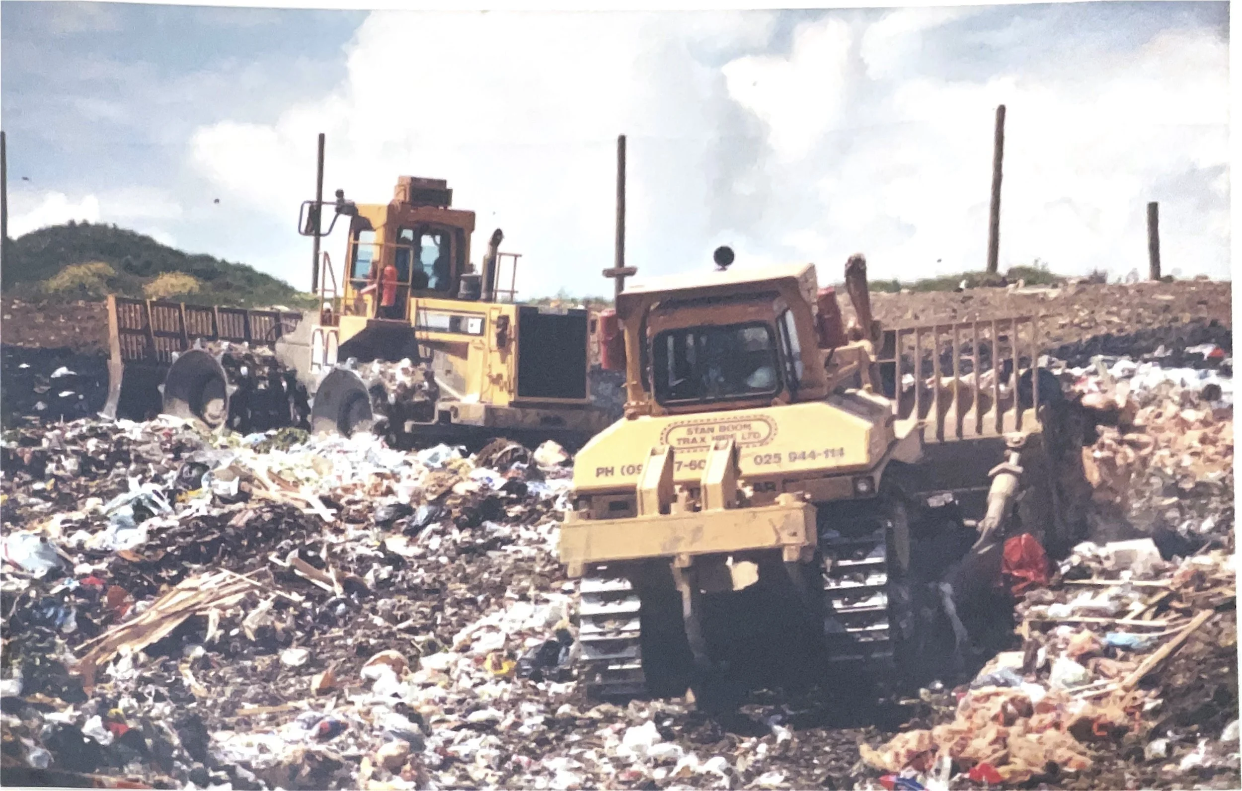 Two bulldozers working on a landfill site with piles of trash and debris, with a partly cloudy sky in the background.