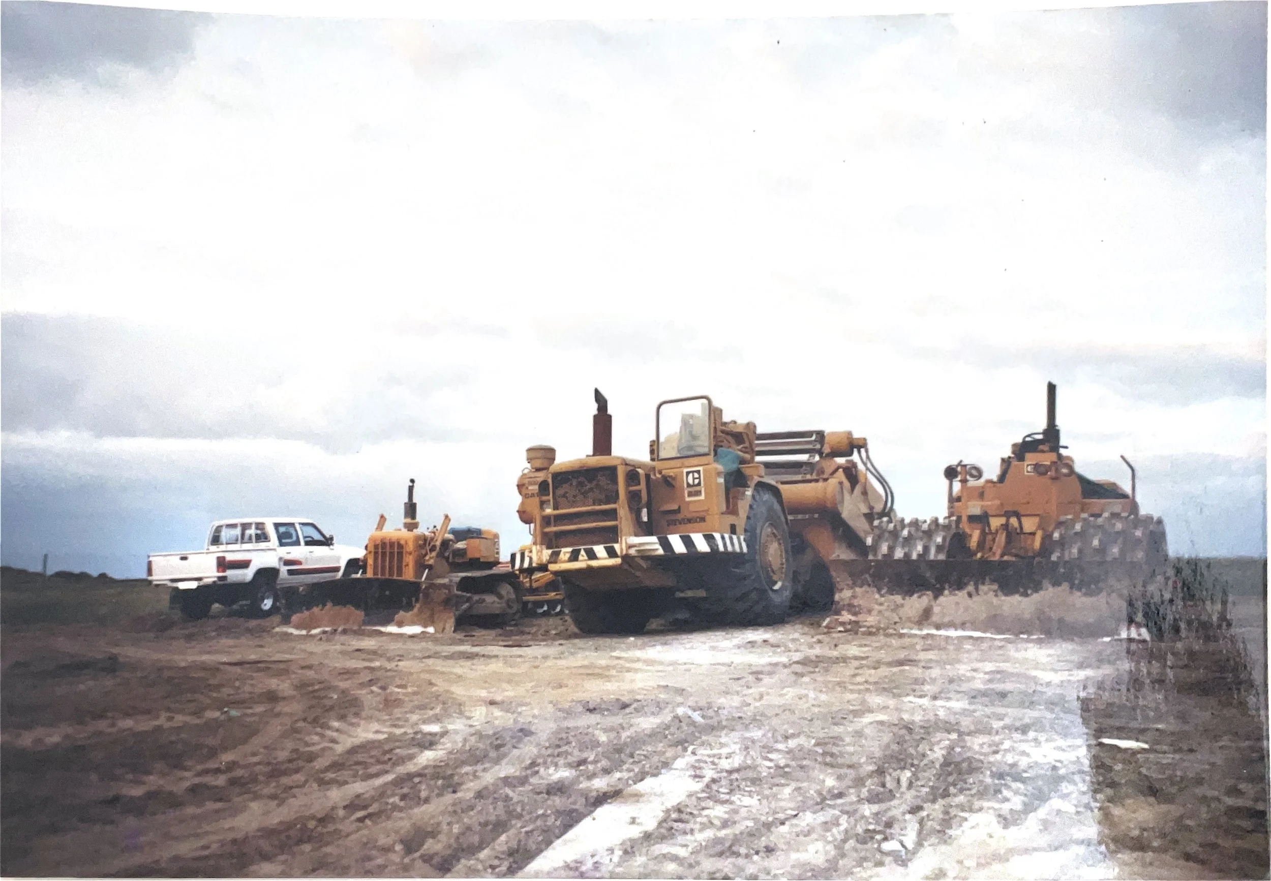 A construction site showing a large yellow bulldozer and a grader working on uneven, muddy ground with water puddles, and a white pickup truck parked on the left, under a cloudy sky.