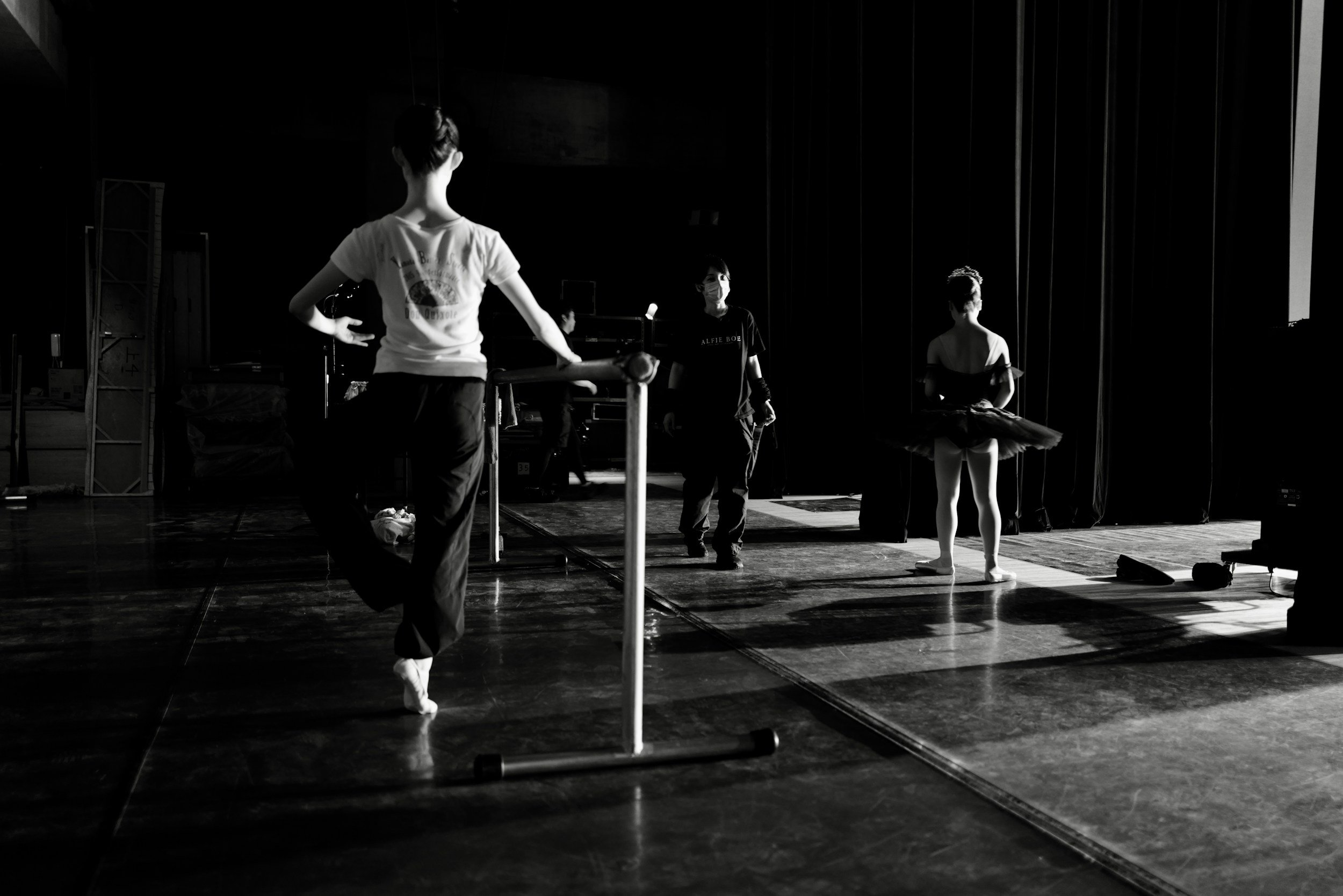 Black and white photo of ballet dancers on stage, with one dancer in front and others in the background, some wearing tutus and masks.