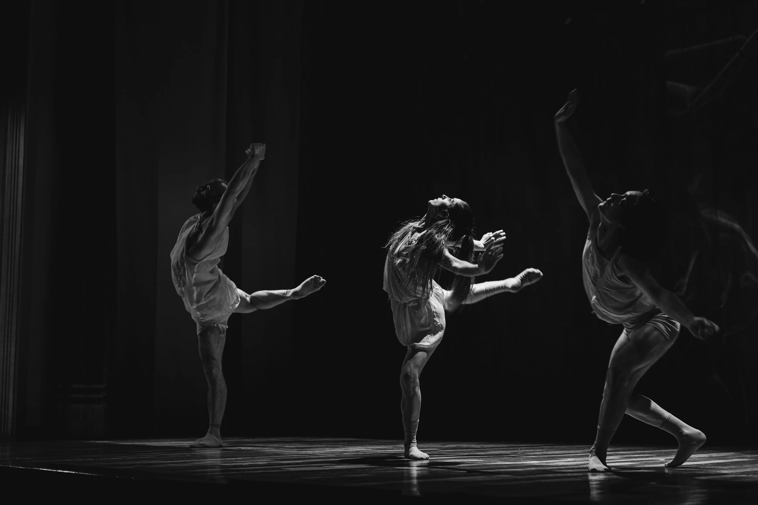 A group of dancers performing on stage in a dark setting, with some dancers standing on one leg and reaching upward, and others in mid-movement with arms and legs extended.