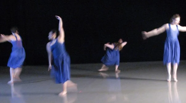 Four dancers in blue dresses performing on stage with a dark background.
