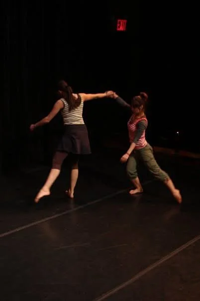 Two women holding hands and dancing on a stage with a dark background and an illuminated red exit sign.