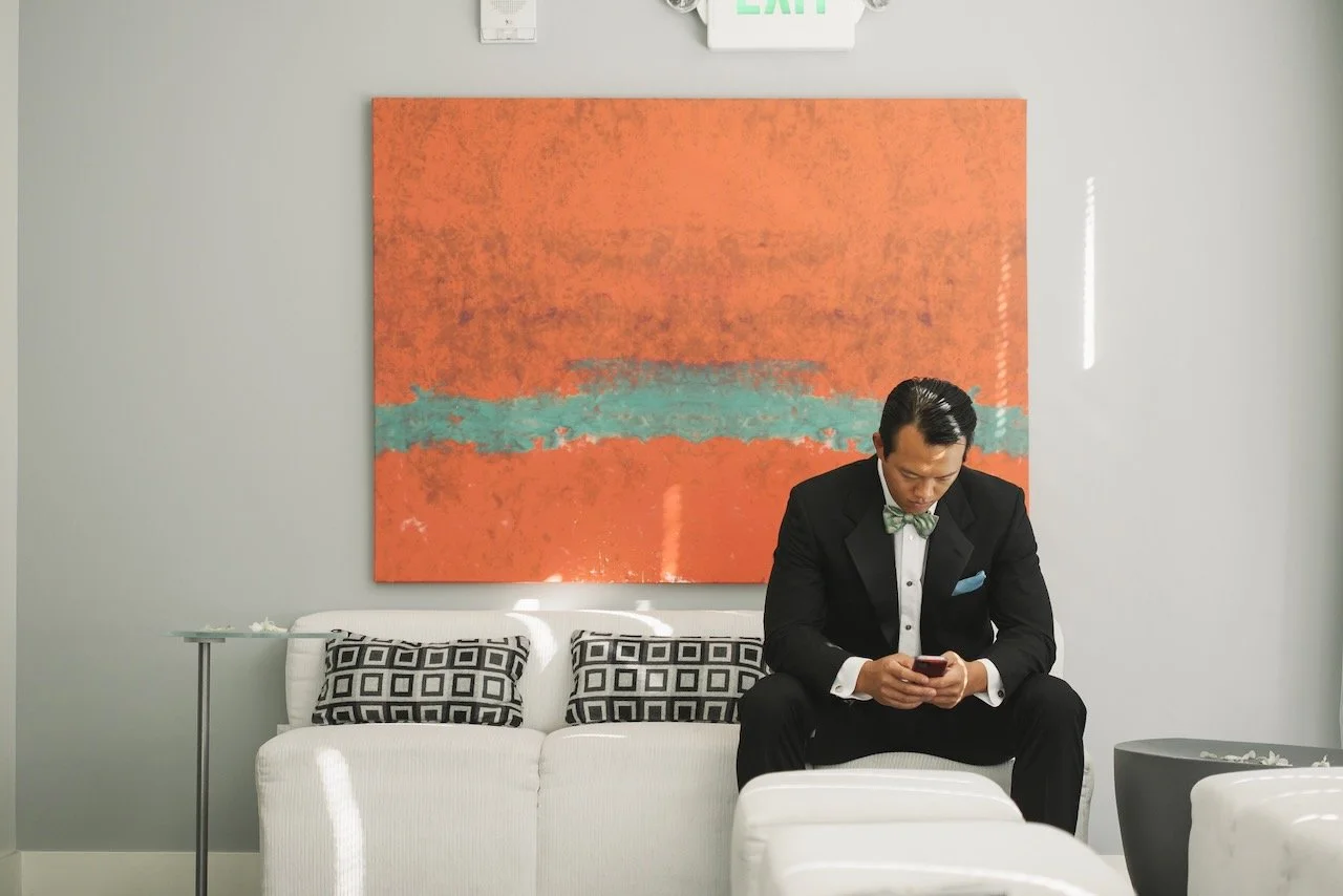 A man in a formal tuxedo with a bow tie sitting on a white sofa, looking at his phone in a modern space with a large orange and green abstract painting behind him.