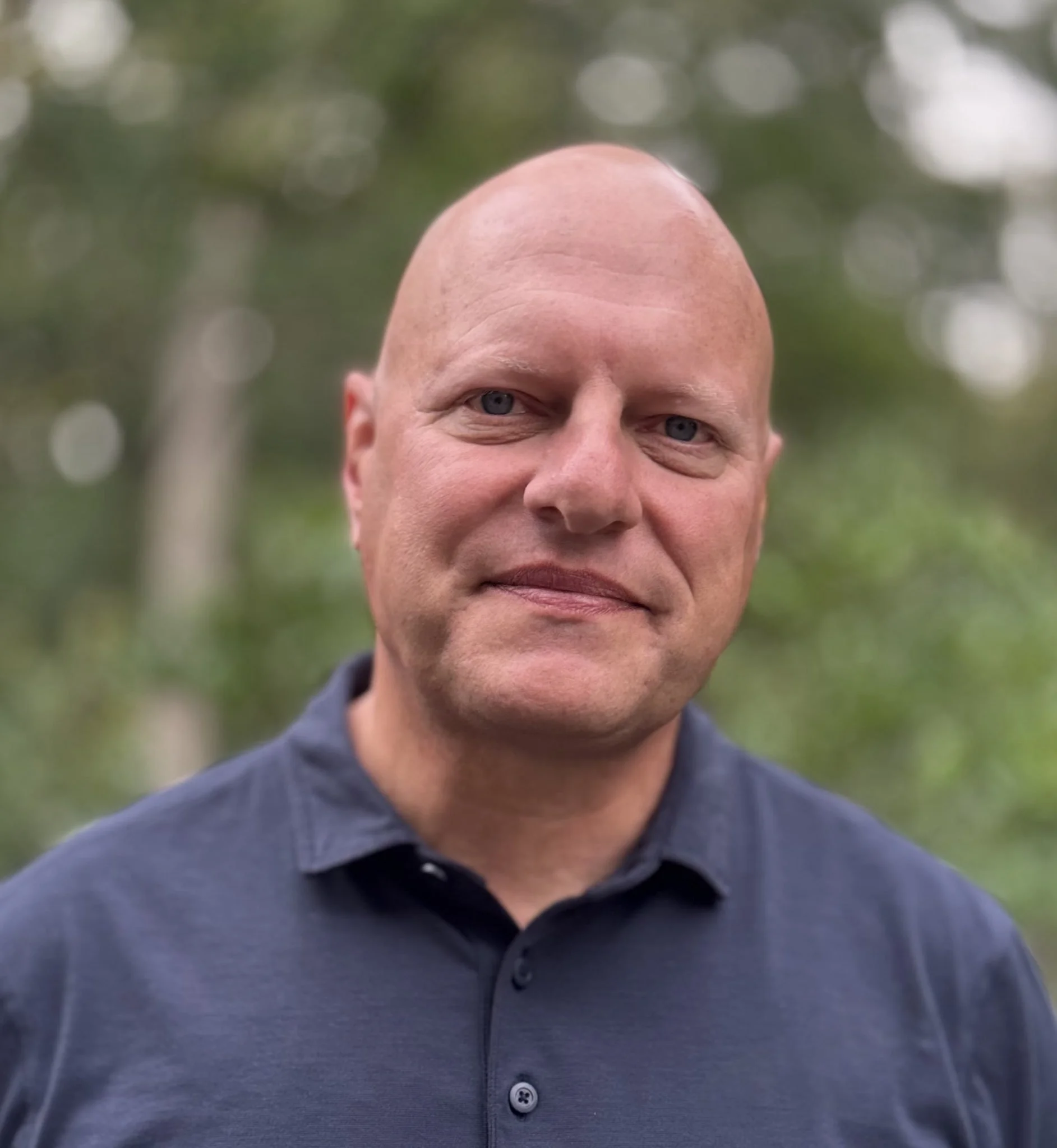 Close-up portrait of a confident, middle-aged man with a shaved head, blue eyes, and a slight smile, wearing a navy blue collared shirt, with a blurred natural green background.