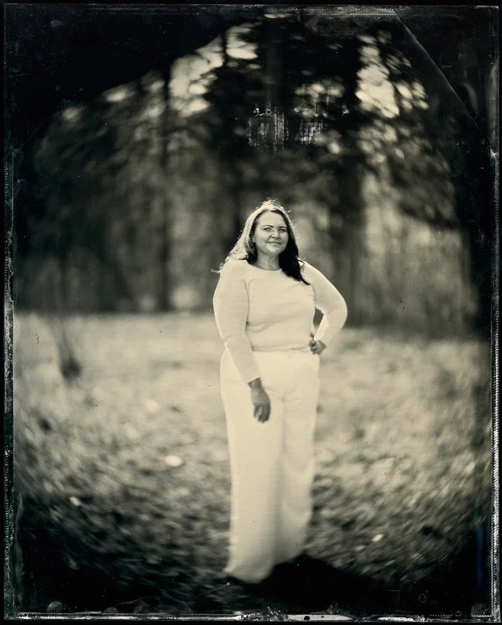 Black and white photograph of a woman standing outdoors on a dirt path surrounded by trees, with her hand on her hip and smiling at the camera.