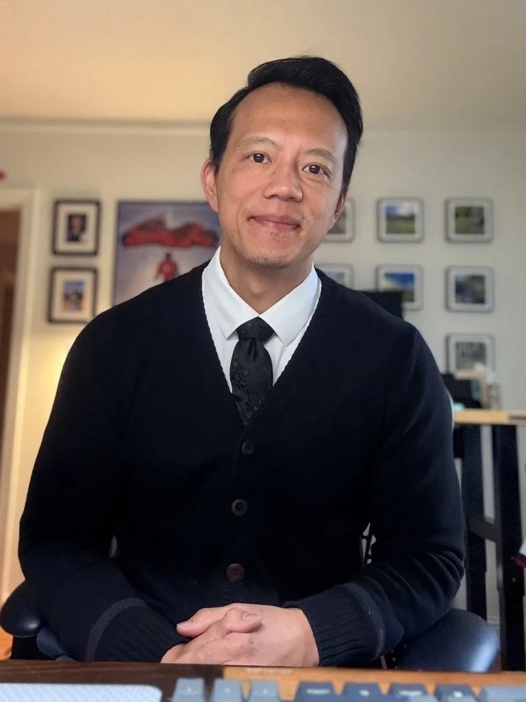 A smiling man with black hair, wearing a white shirt, black tie, and black cardigan, seated indoors with a wall of framed pictures behind him.