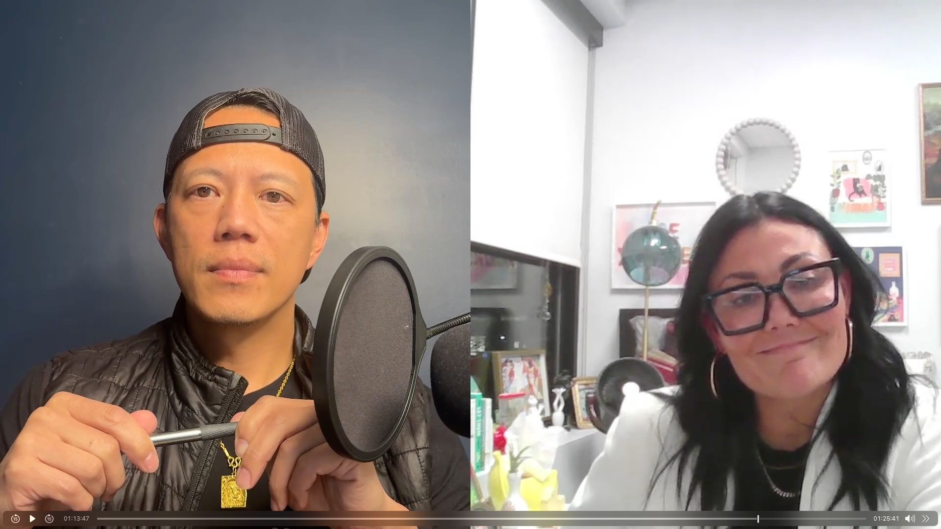 A man wearing a black baseball cap backwards and a black jacket holding a microphone, and a woman with black hair, glasses, and a white blazer smiling in a room with colorful artwork on the wall.