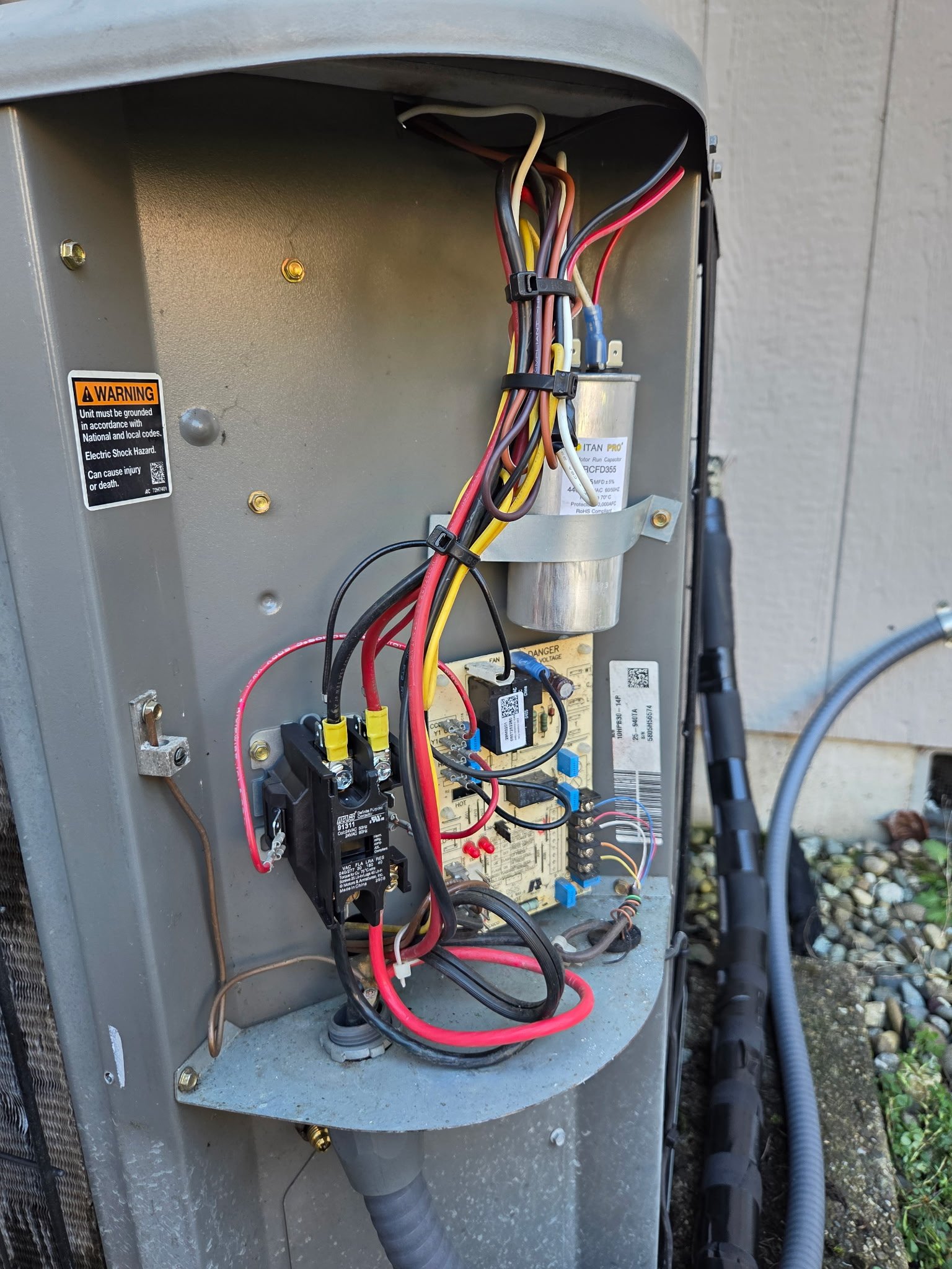 Open HVAC unit with wiring, circuit board, relay, and capacitor inside, mounted outside on a concrete surface.