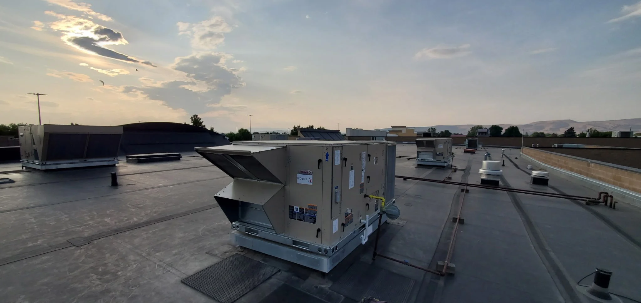 Rooftop HVAC units and ventilation systems under a partly cloudy sky with the sun setting behind clouds, and hills in the distance.