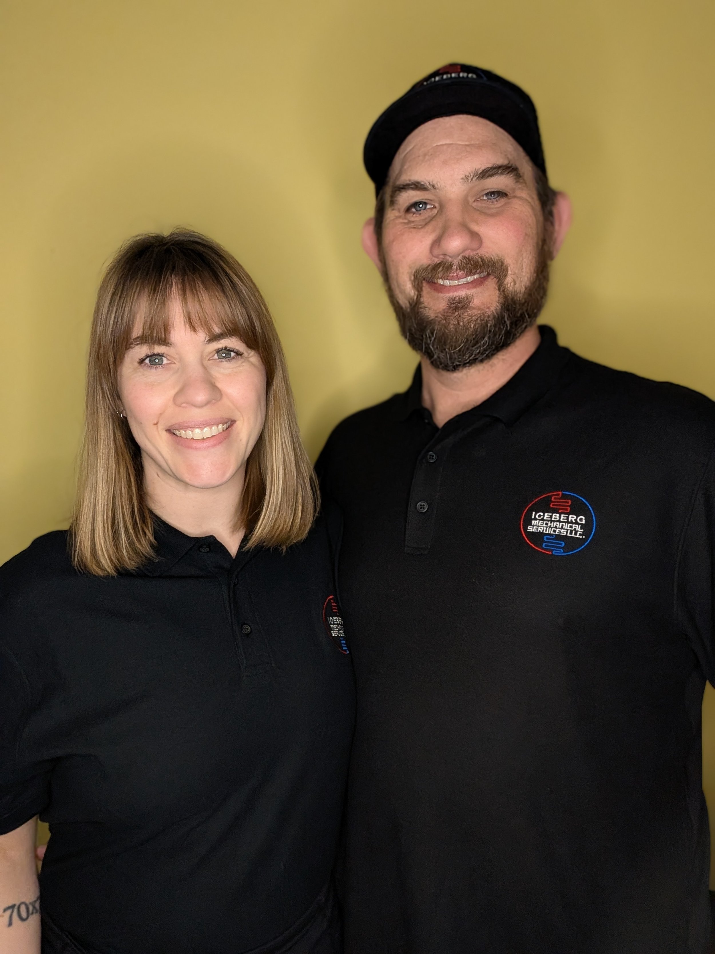 A woman and a man standing together, both wearing black polo shirts with a logo that says Iceberg Mechanical Services LLC, smiling in front of a yellow background.