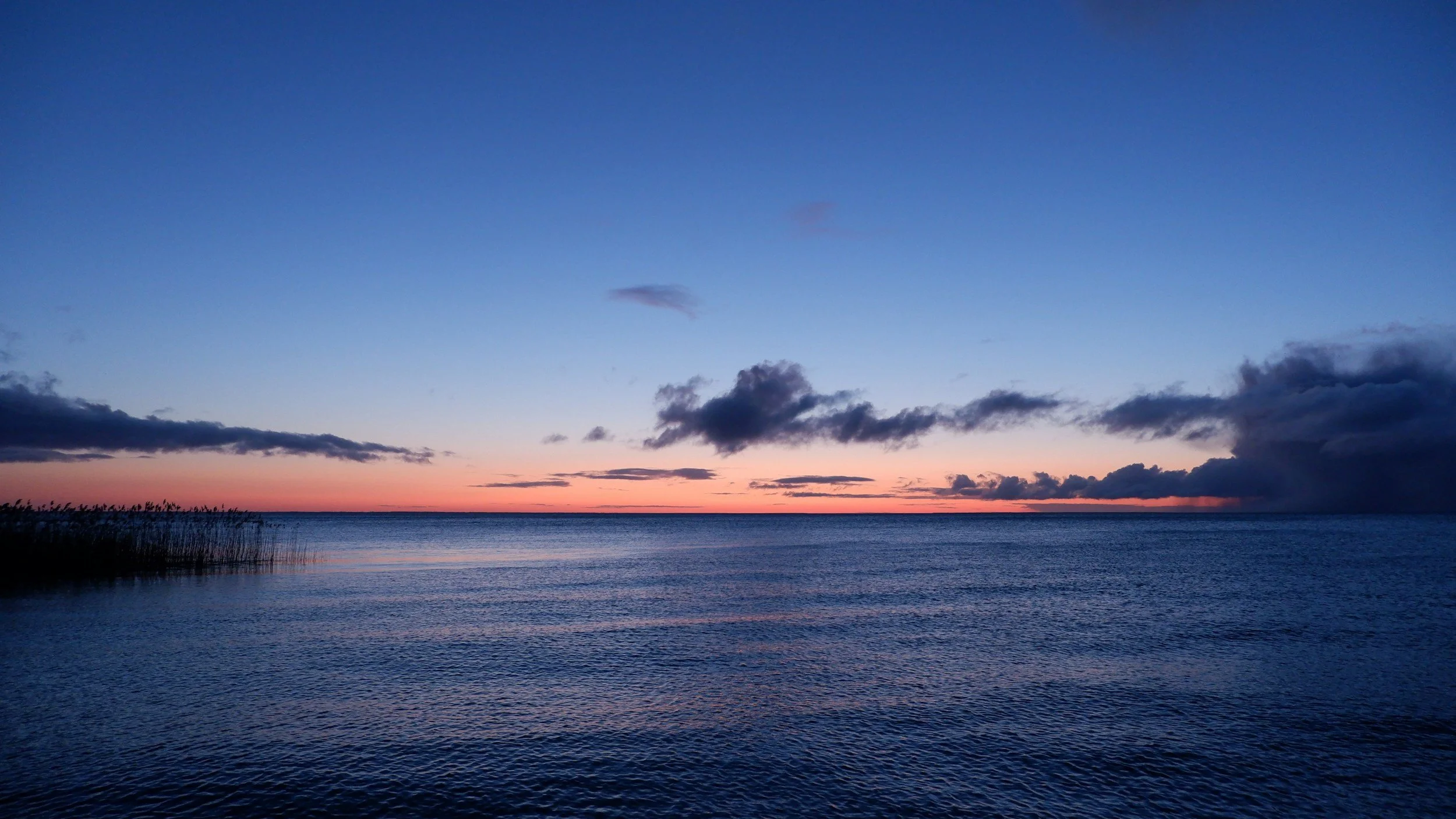 A peaceful scene of a body of water at sunset, with a colorful sky featuring orange, pink, and blue hues. There are some clouds on the horizon and plants in the foreground.