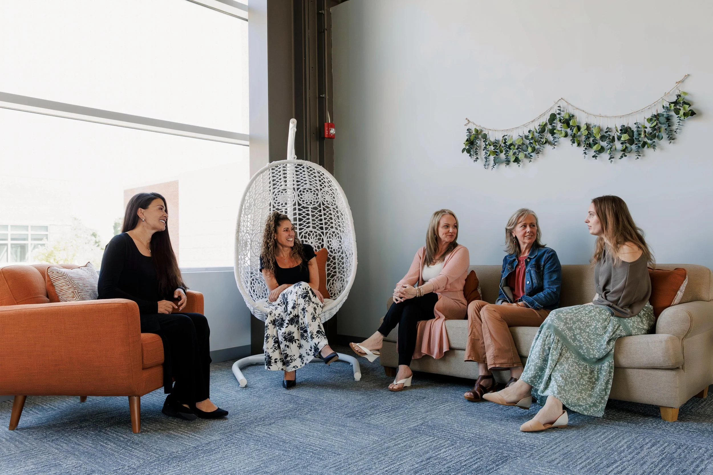 Group of five women sitting in a living room, smiling and talking, with a hanging white chair, green leaf wreath wall decoration, large window, and orange and beige sofas.