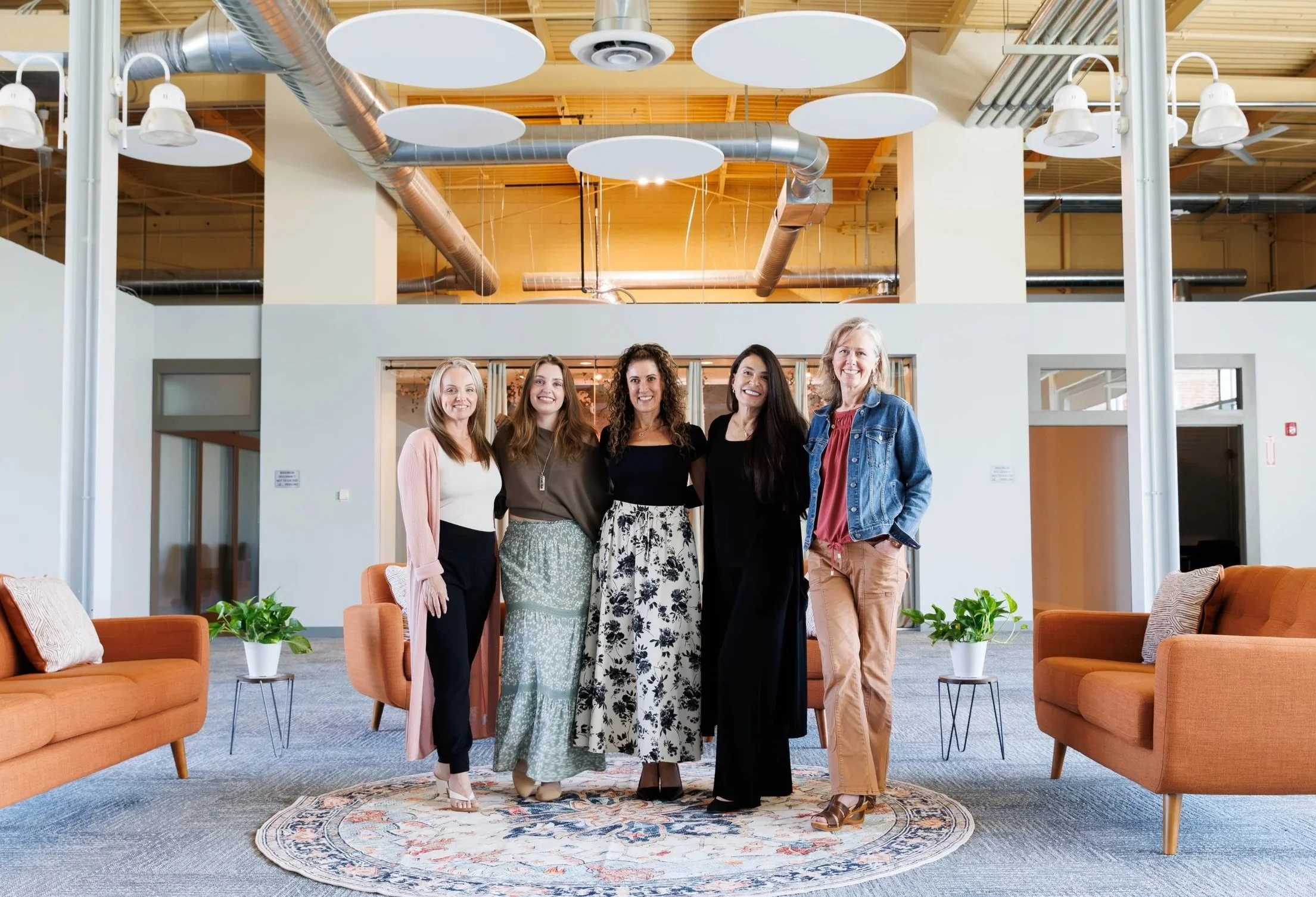 Five women standing together in an office lounge area, smiling at the camera, with orange sofas and potted plants in the foreground and modern industrial ceiling with ducts above.